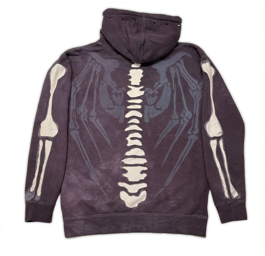 A dark plum-purple hoodie shown from the back, featuring a large graphic print of a human skeleton spine running down the center with skeletal arms and wings extending out on both sides.