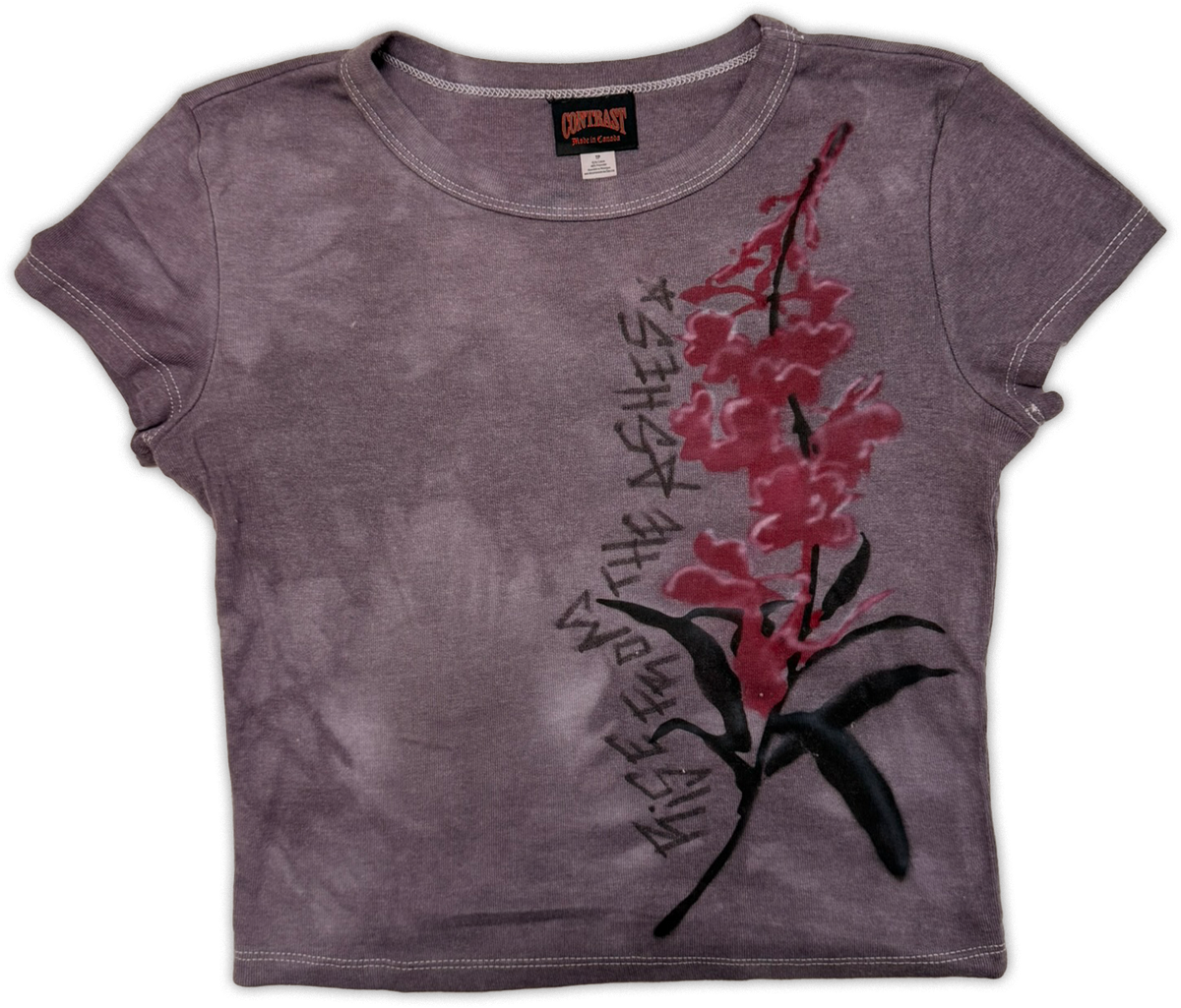 A short-sleeve crop top t-shirt in a washed, faded purple-gray color, featuring a graphic print of a red and pink flower with black leaves and stylized text running vertically alongside it.