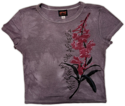A short-sleeve crop top t-shirt in a washed, faded purple-gray color, featuring a graphic print of a red and pink flower with black leaves and stylized text running vertically alongside it.