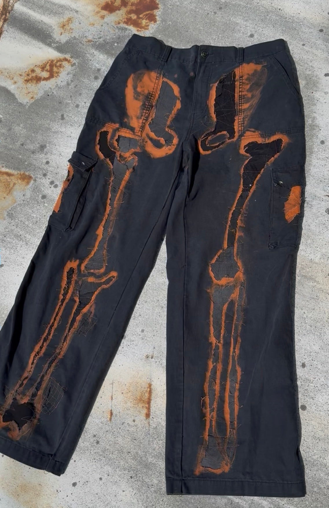 Black cargo pants with a distressed orange-brown bone or skeletal graphic printed down the front legs, laid flat on a weathered concrete surface.
