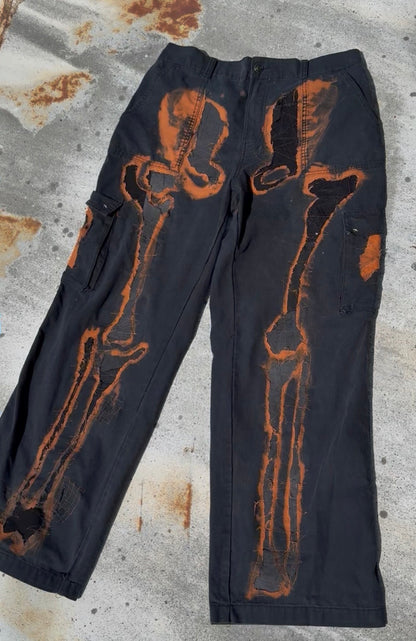 Black cargo pants with a distressed orange-brown bone or skeletal graphic printed down the front legs, laid flat on a weathered concrete surface.