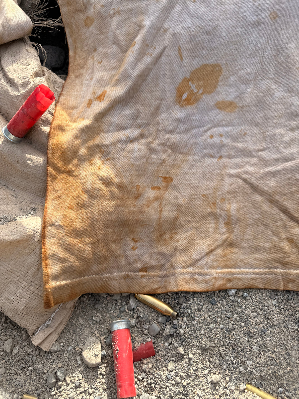 A close-up of a stained beige fabric laid on gravel, with red shotgun shells and brass casings scattered nearby.