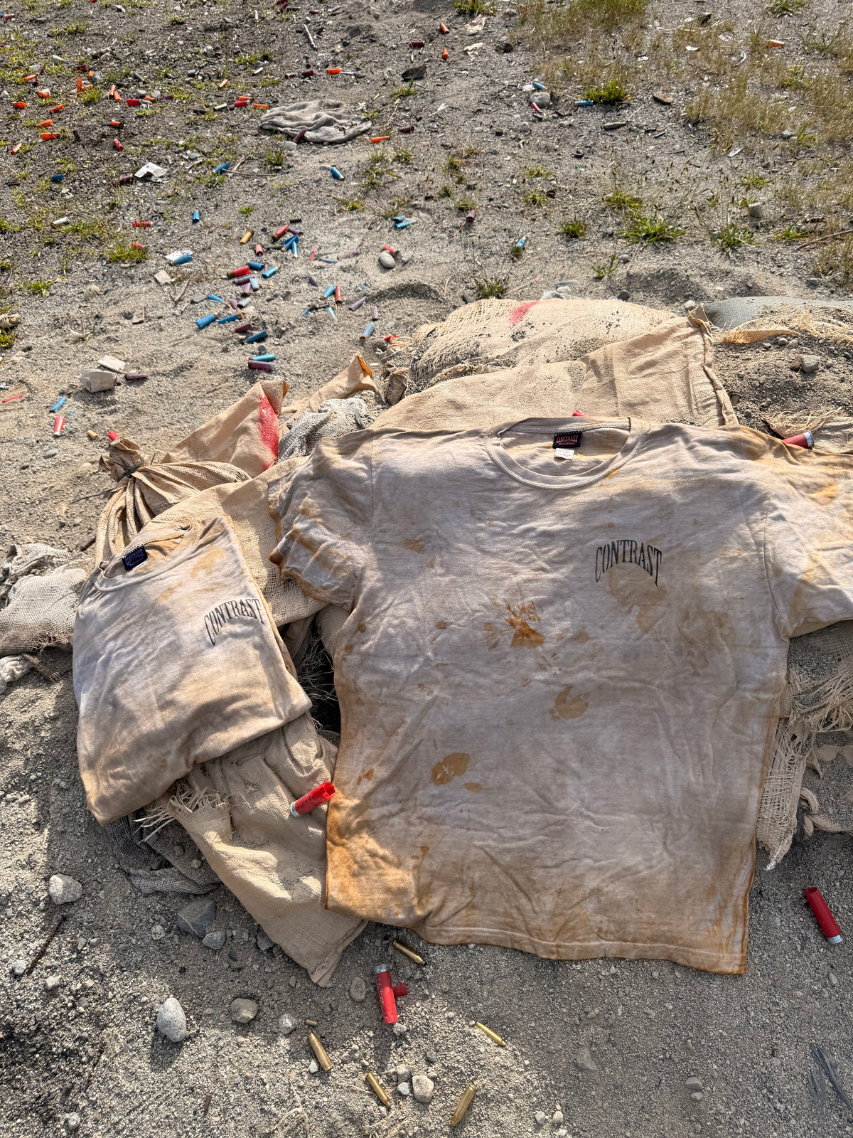 A stained beige t-shirt and matching sweatshirt laid out on sandy ground outdoors, with scattered bullet casings visible around them.