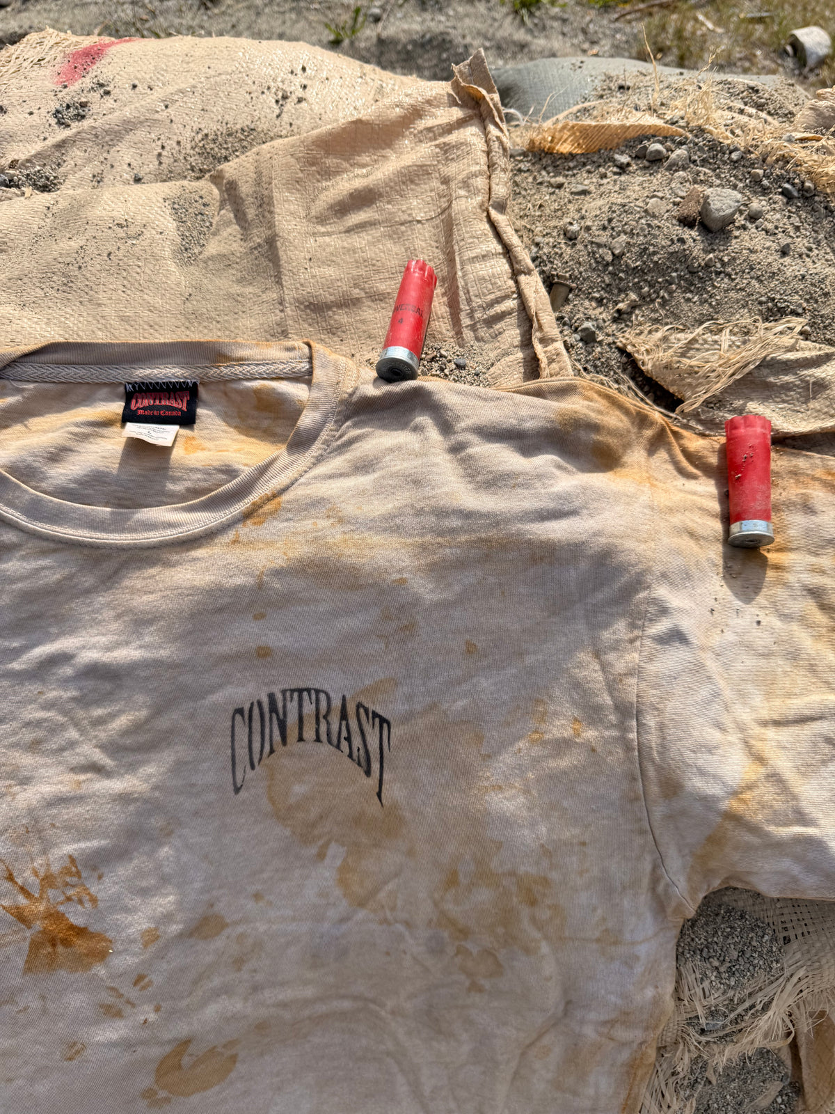 A dirty off-white t-shirt with the word 'CONTRAST' printed in black on the chest, laid flat on a sandy surface with two shotgun shells placed on the shoulders.