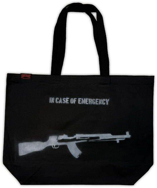 A black canvas tote bag with black handles, featuring the text 'IN CASE OF EMERGENCY' and a gray silhouette of a submachine gun on the front. A small red tag is visible on the left side.