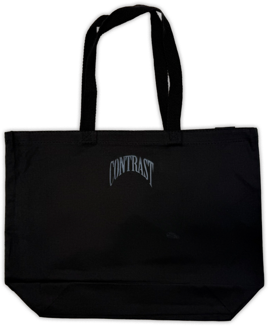 A black canvas tote bag with two shoulder straps and a stylized gray 'CONTRAST' logo printed on the front, shown against a plain white background.