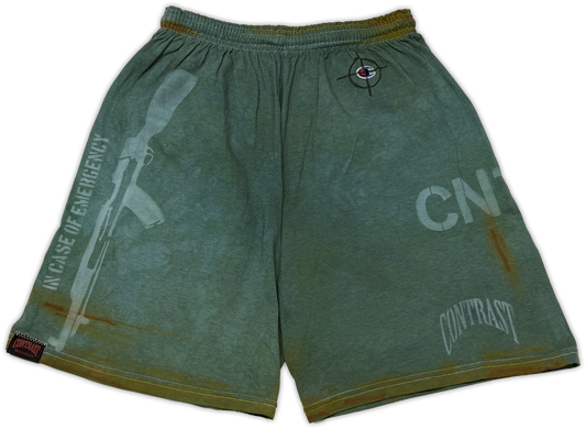 A pair of distressed sage green athletic shorts with tactical-style graphics, including a rifle silhouette, 'IN CASE OF EMERGENCY' text, and 'CONTRAST' branding.