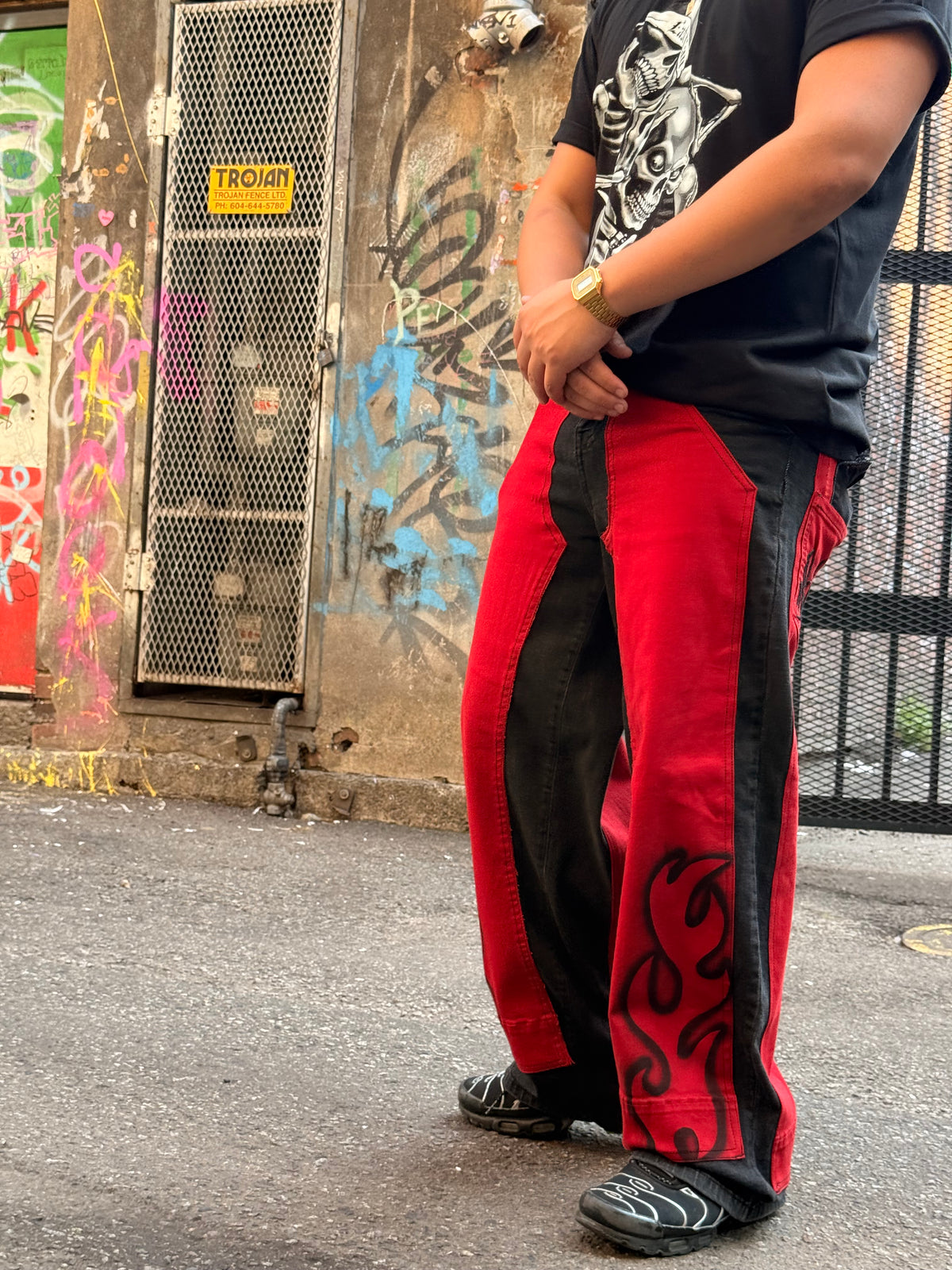 A person wearing red and black wide-leg pants with a black graphic t-shirt, standing on asphalt in front of a graffiti-covered wall.