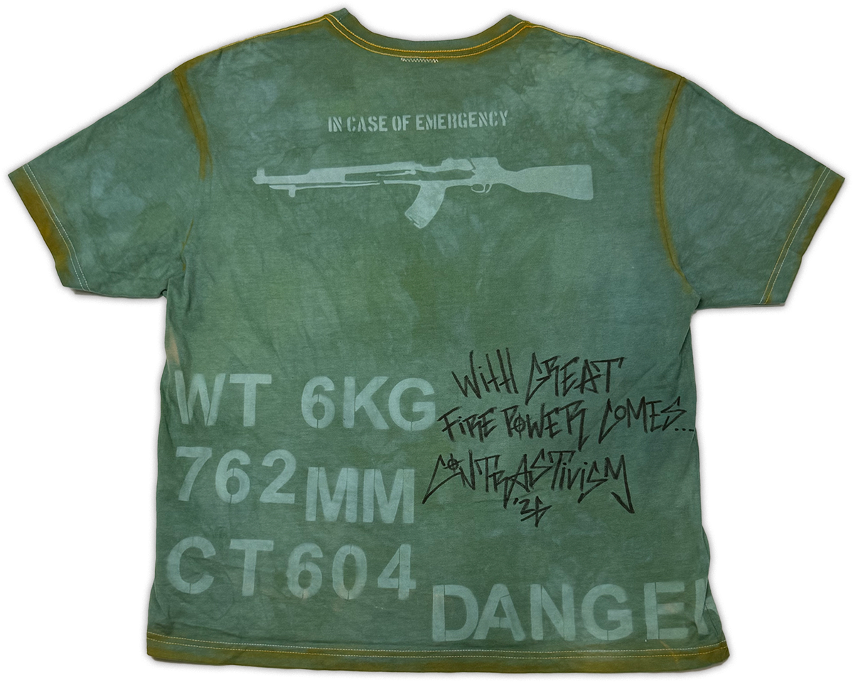 A green distressed short-sleeve t-shirt with military-style graphics and text, including a rifle silhouette, 'IN CASE OF EMERGENCY' at the neck, and 'DANGER' with handwritten-style text on the back.