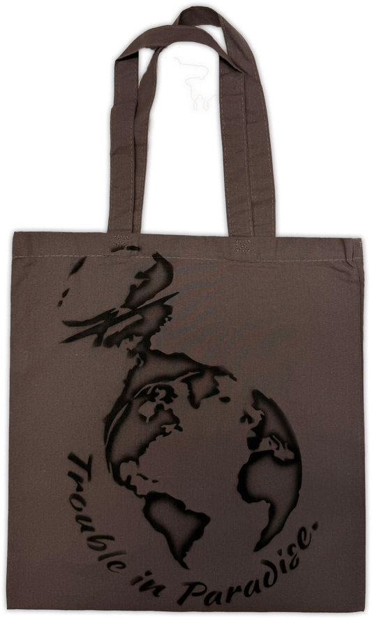 A dark brown canvas tote bag with long handles, featuring a black graphic of a globe being nuked and the text 'Trouble in Paradise.' printed on the front, set against a plain white background.